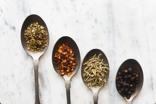 Four silver spoons displaying different dried herbs and spices in a row.